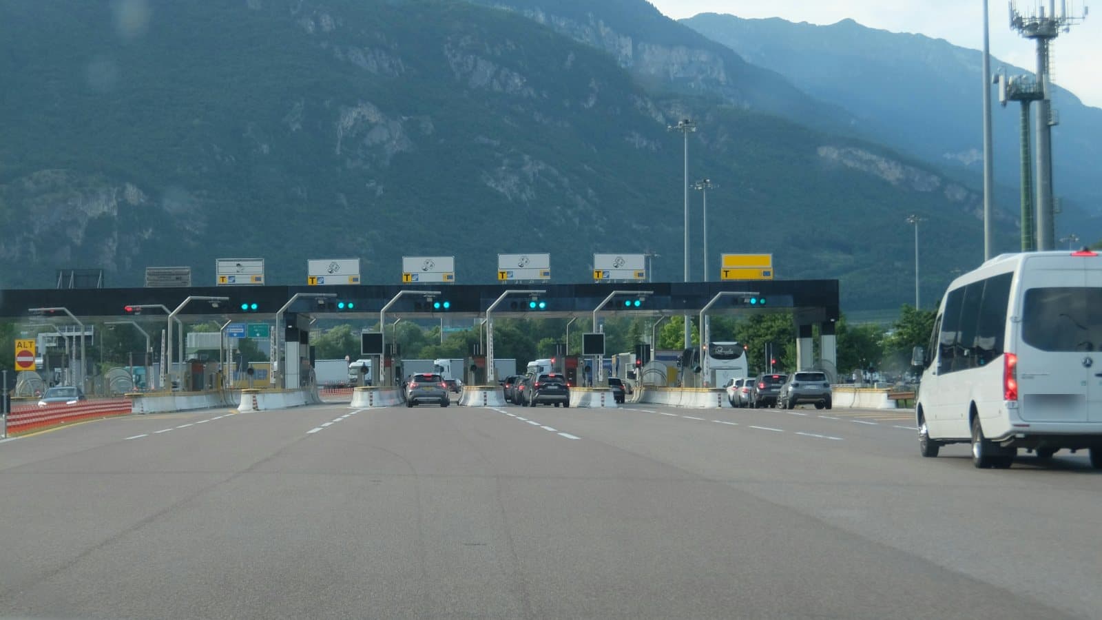 Cars and trucks at a motorway toll plaza in Italy, with mountains behind